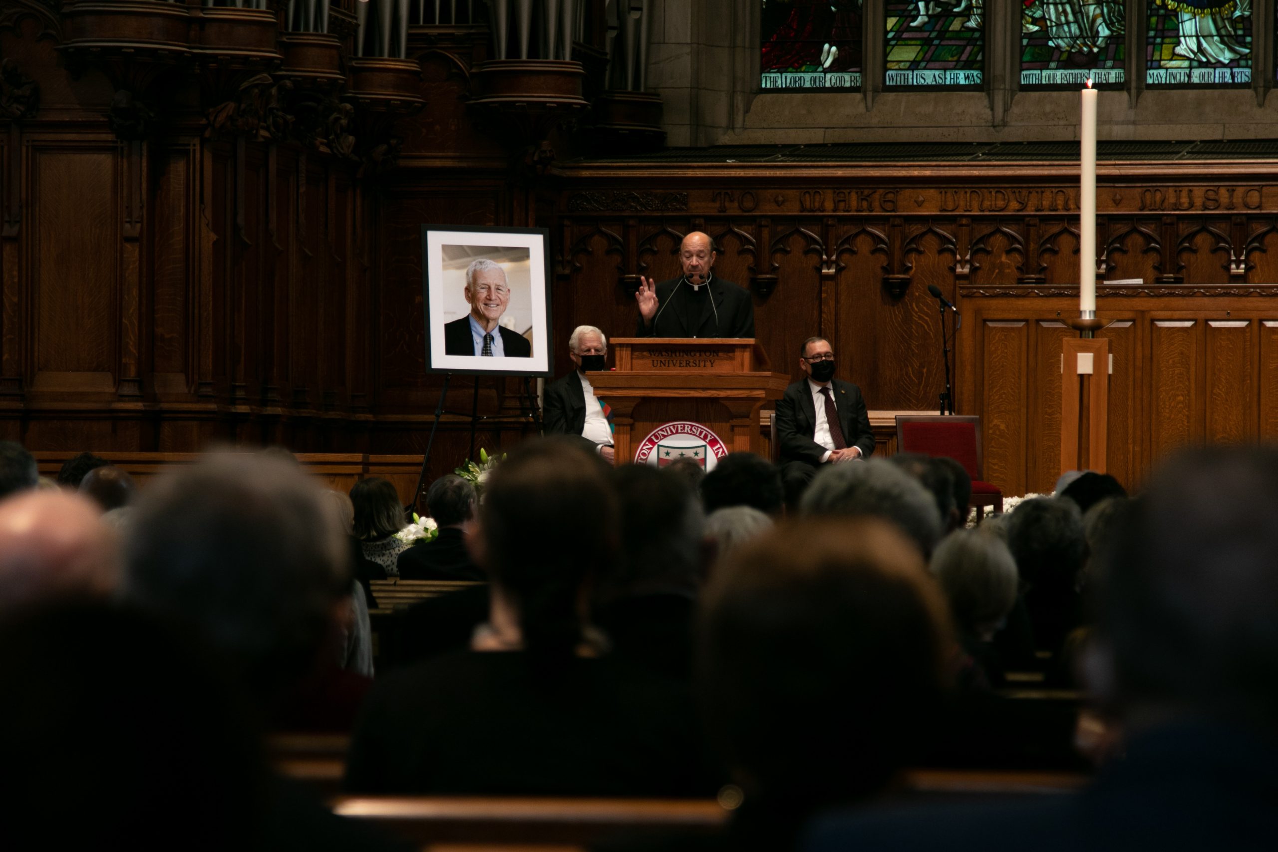 The heads of a crowd of people are visible and in the distance a man in black robes stands at a lectern. A poster with the face of a man in a suit and tie stands to the left of the lectern.