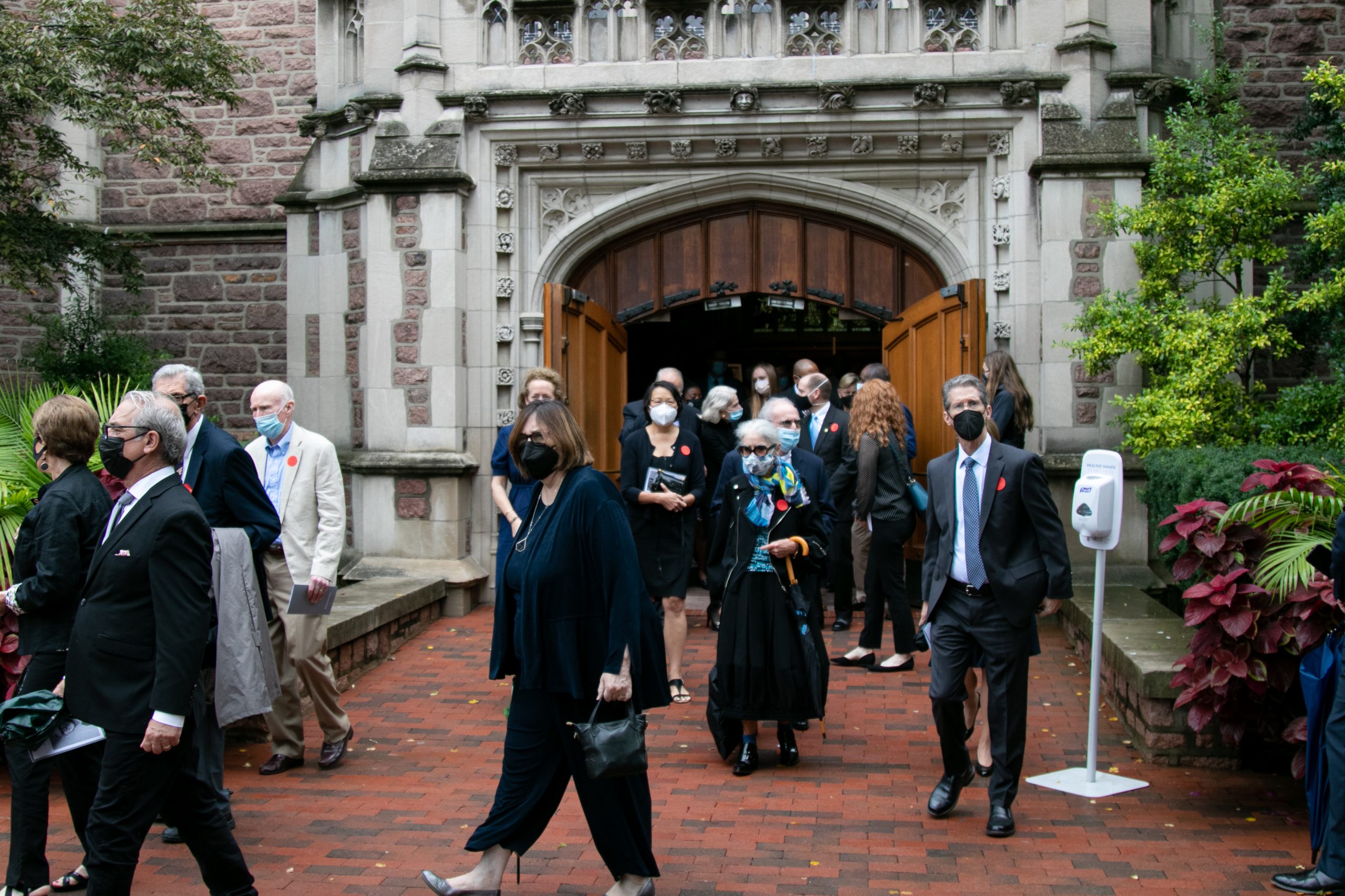 People in formal attire walk on brick walkways outside of a building with an arch and wooden doors.