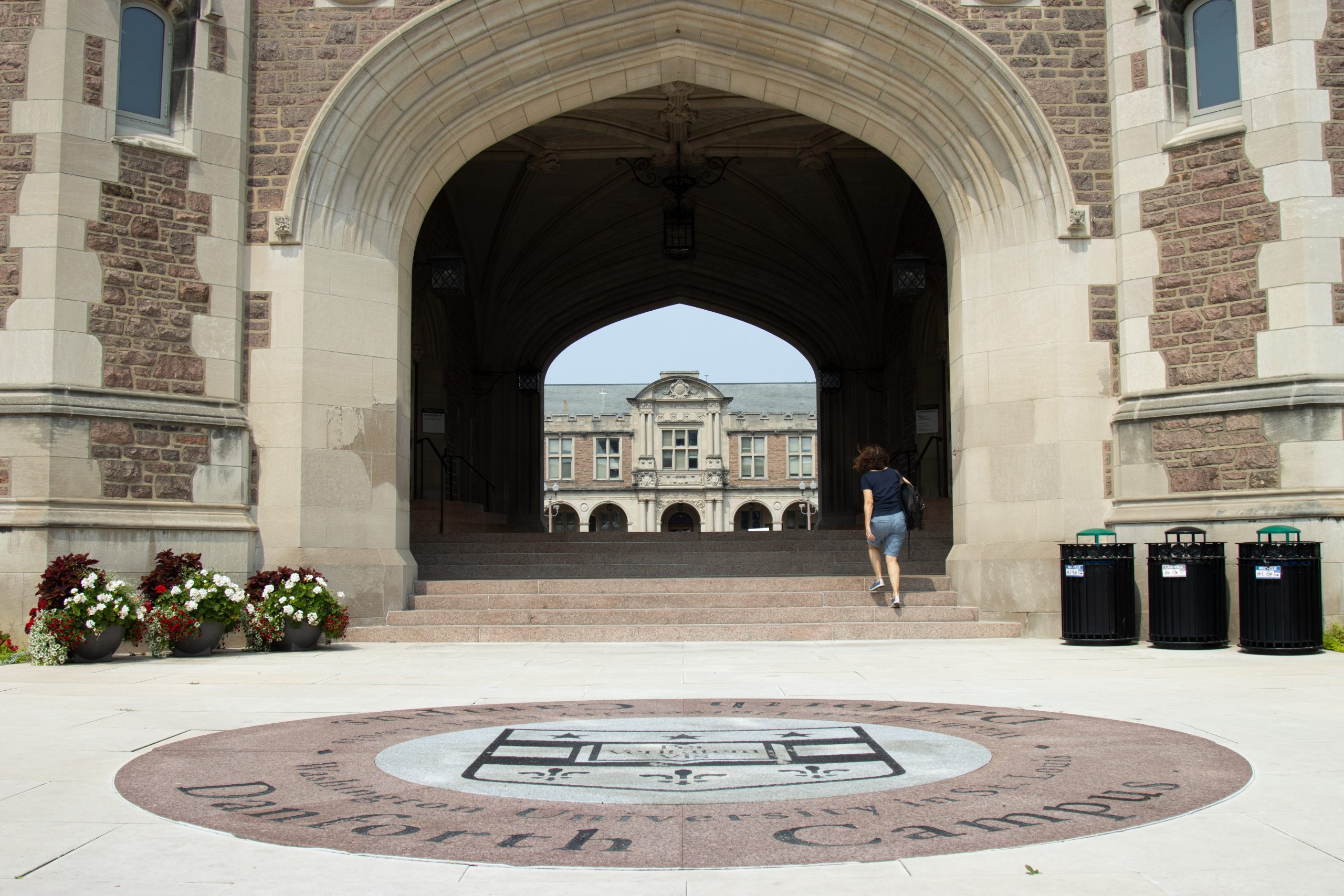 The Washington University seal sits in front of stairs leading under an arch. A person walks up the right side of the stairs.