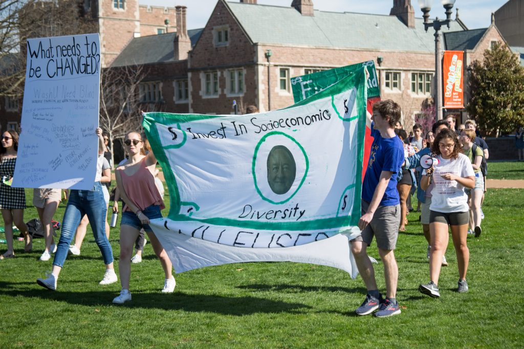 Two students, one on the left wearing a pink shirt and one on the right wearing a blue shirt, hold a large white banner with green trim that reads "Invest in Socioeconomic Diversity" and features coloring that makes it look like a dollar bill with former chancellor Mark Wrighton's face in the middle. Other students walk behind the banner on a sunny day with a brick building in the background. The students stand on grass.
