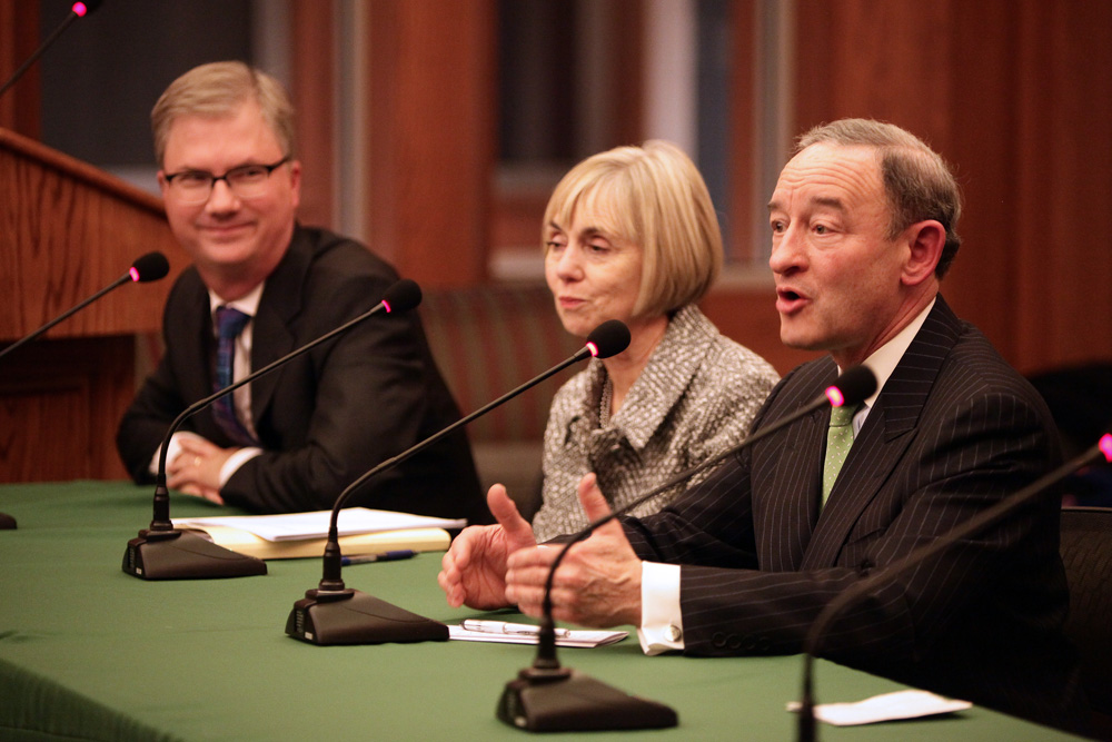 Three people in suits sit in front of microphones atop a green tablecloth. Wood panels and windows are behind them.