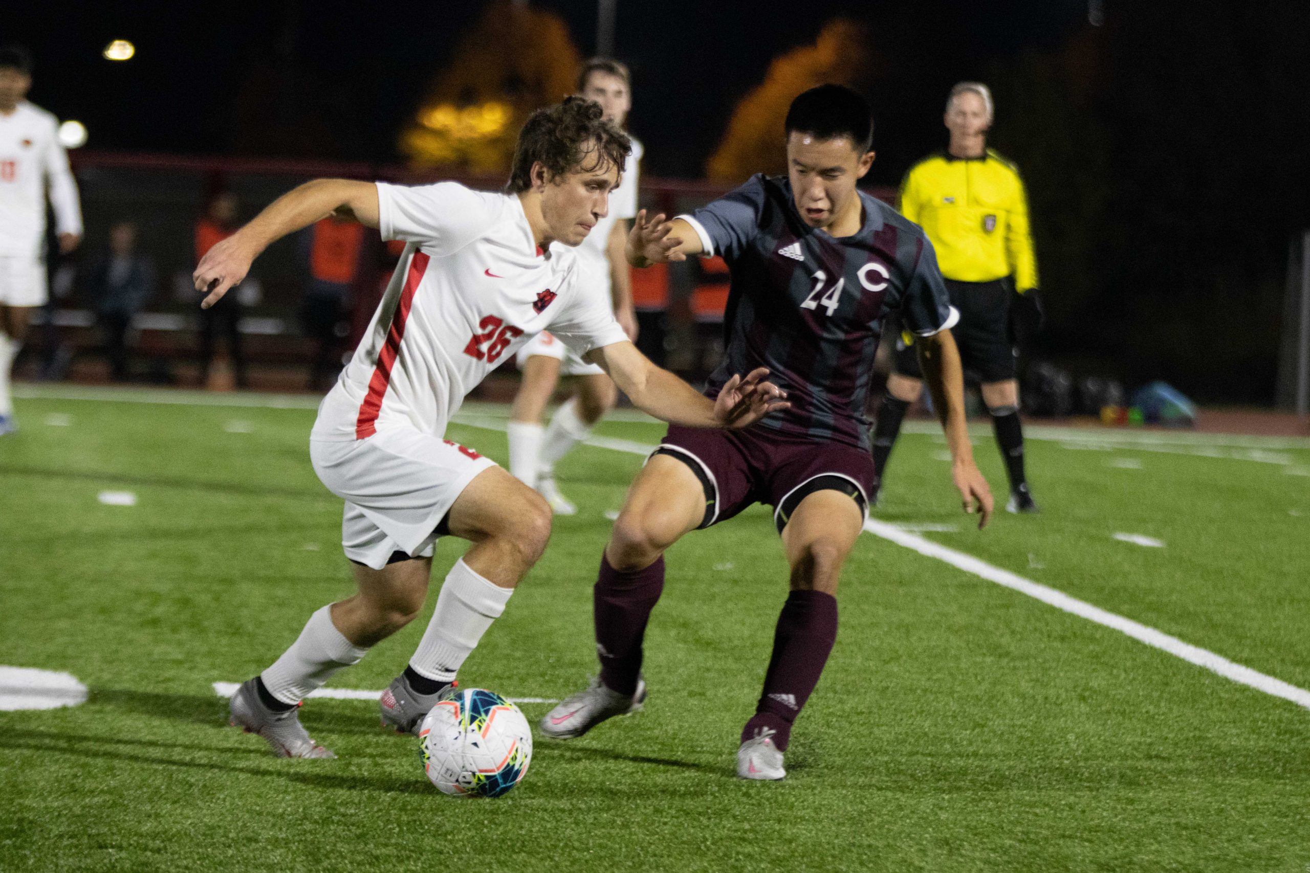 A soccer player in a white jersey with a red stripe down the right side controls the ball with his right foot as a defender in a black jersey attempts to stop him. A referee in a yellow shirt stands in the background.