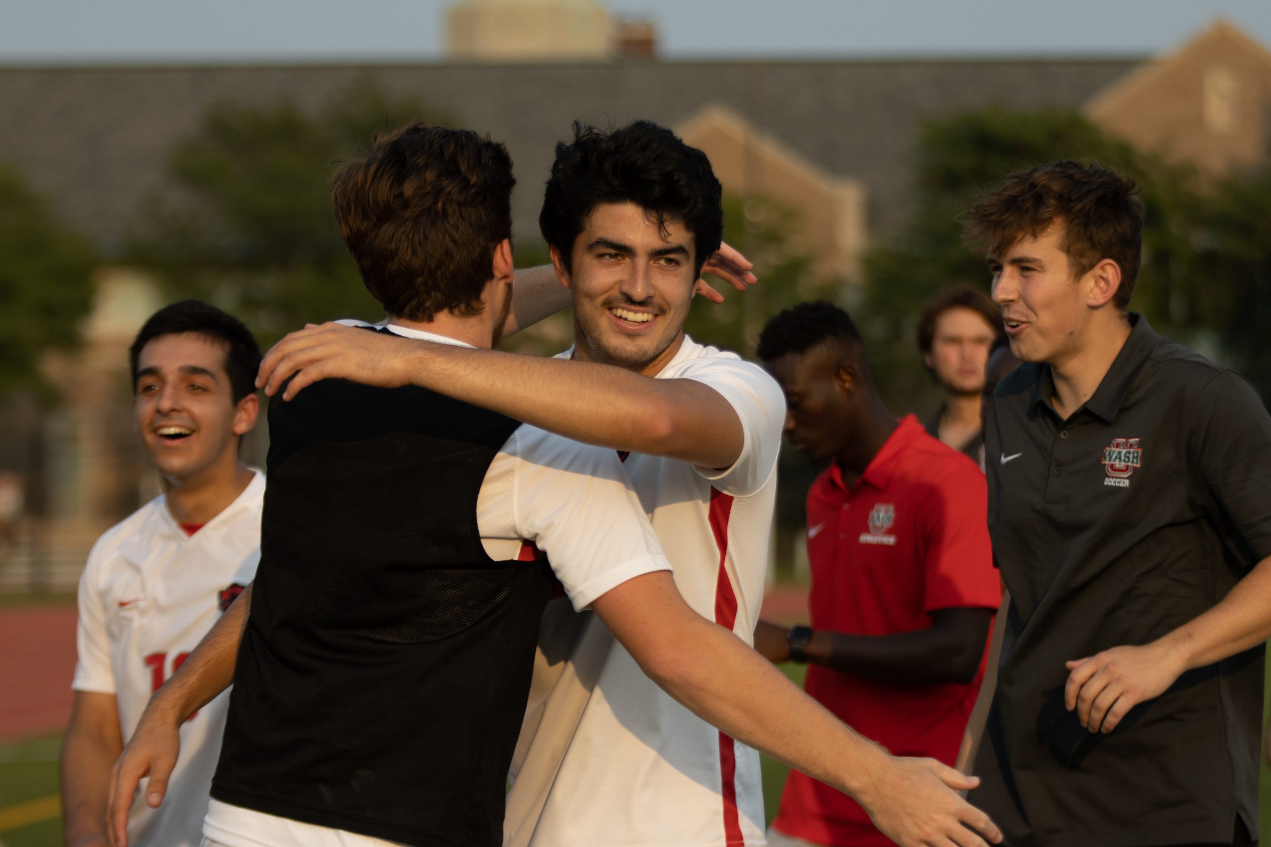 A soccer player in a white jersey places his left arm around a player in a white jersey with a black vest as the two hug. A player in a red shirt stands in the background.