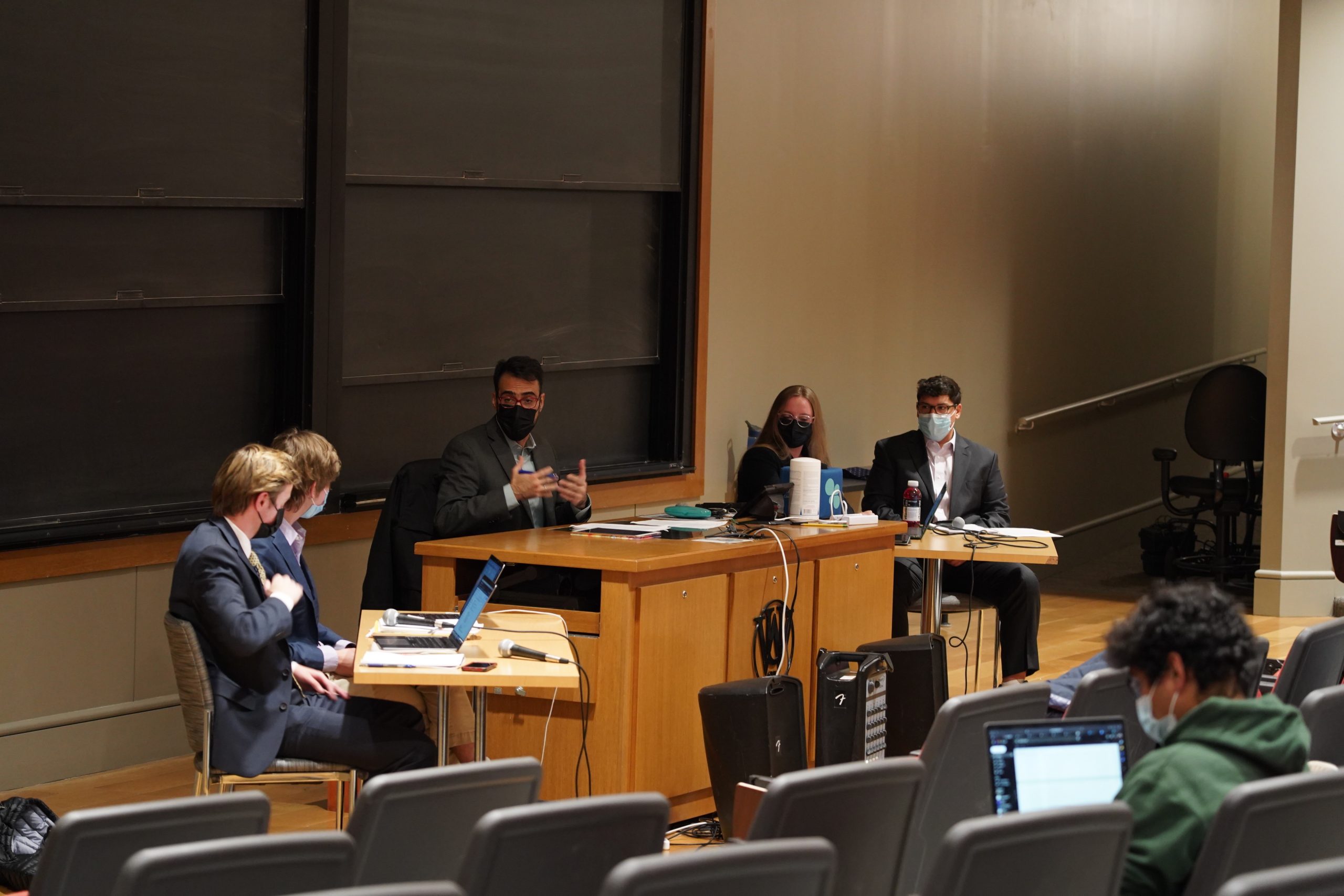 Five masked individuals sit behind a wooden desk with a blackboard behind them. A person with an open laptop screen can be seen sitting in a seat in the foreground