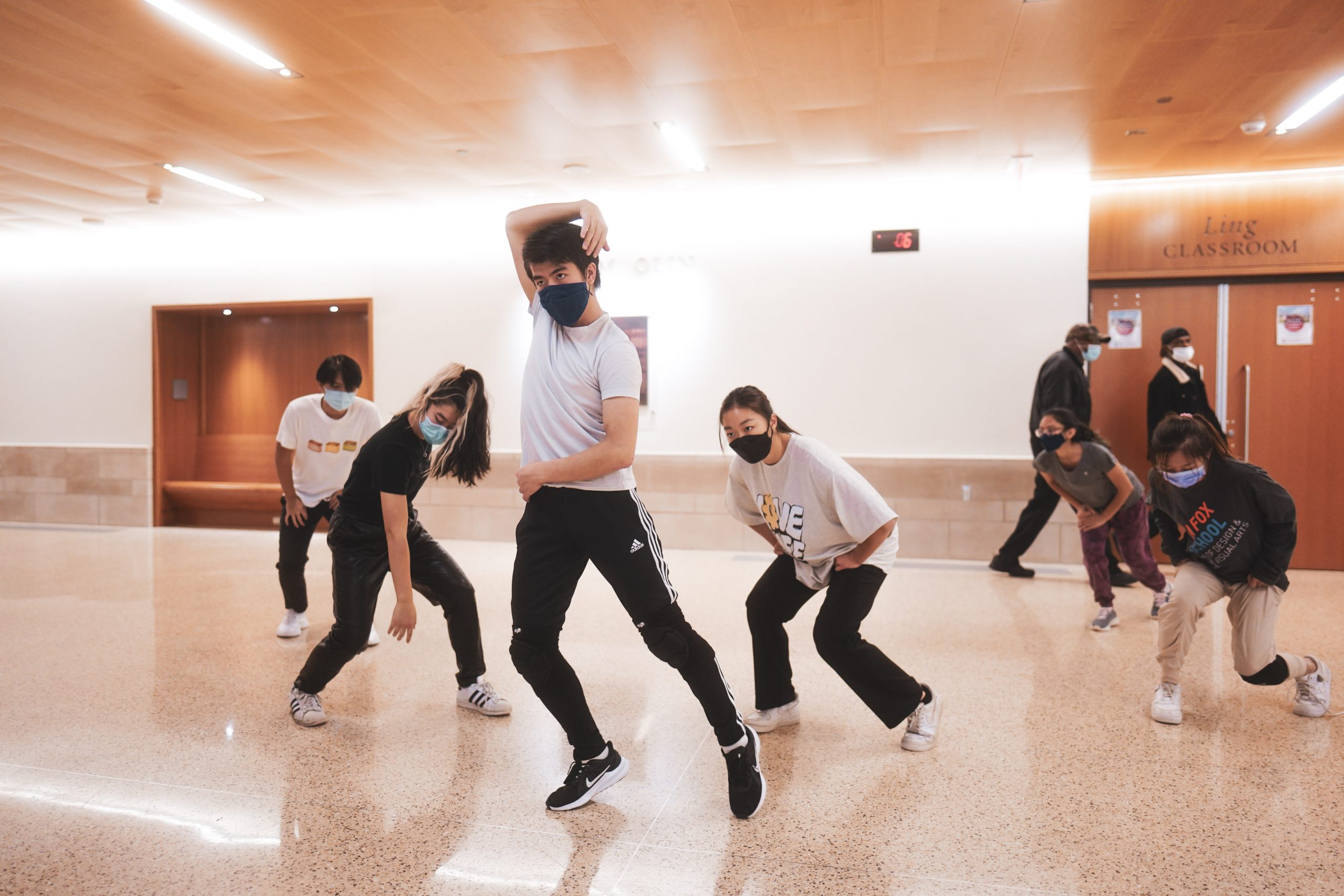 Six masked people in black sweatpants and grey shirts strike various poses in a well lit room