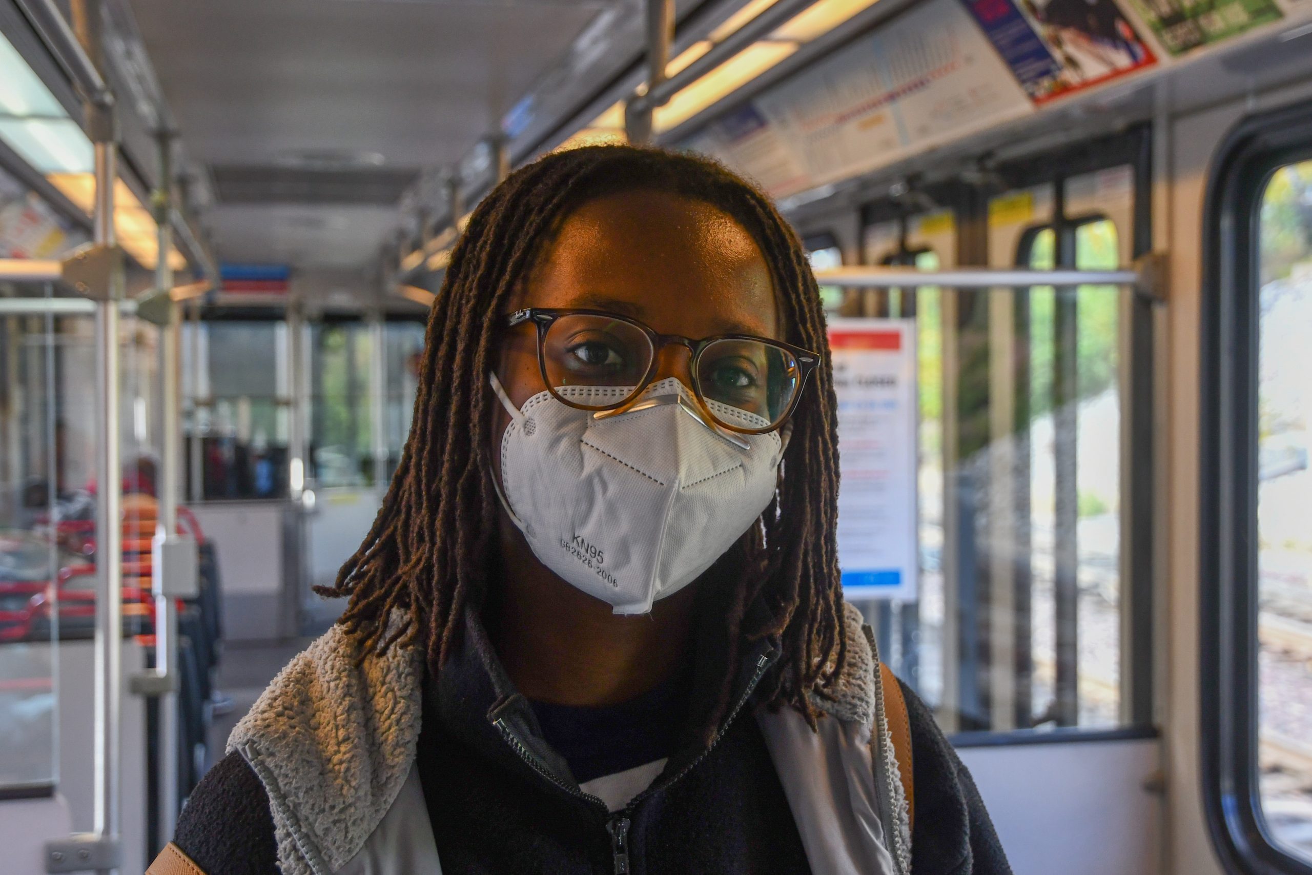 A person with big round glasses, long dark hair and a light blue face mask wears a gray vest as the MetroLink is visible in the background.