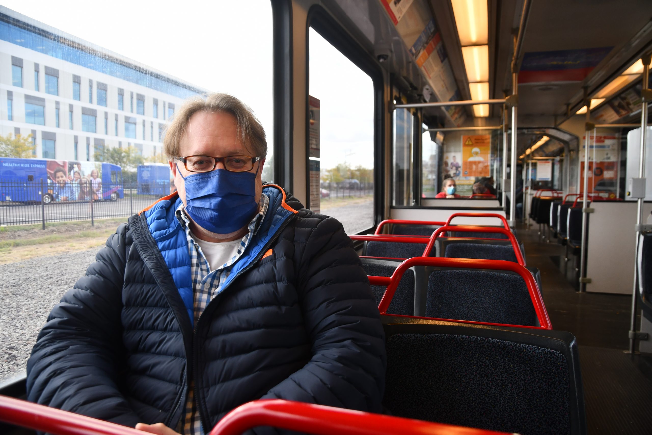 A person wearing a blue and navy jacket sits on red seats on the Metro while wearing a dark blue mask. It is cloudy outside and there is a building out the window.