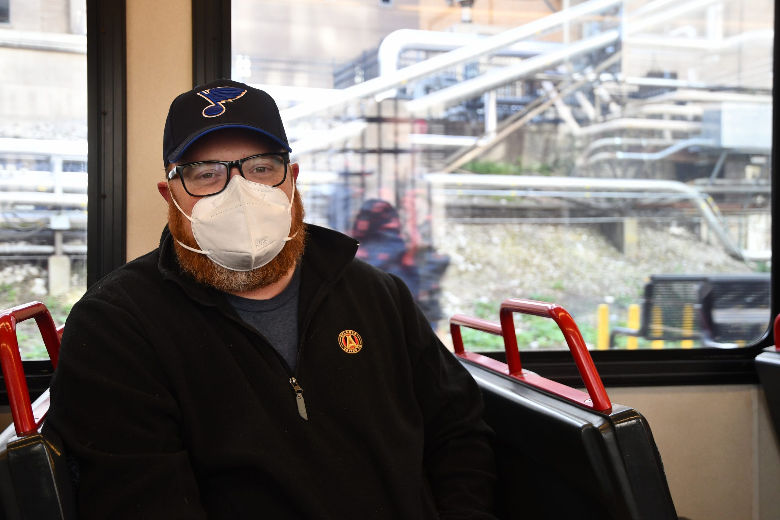A man with a large red beard wears a white mask, a dark fleece and a blue St. Louis Blues hat. 