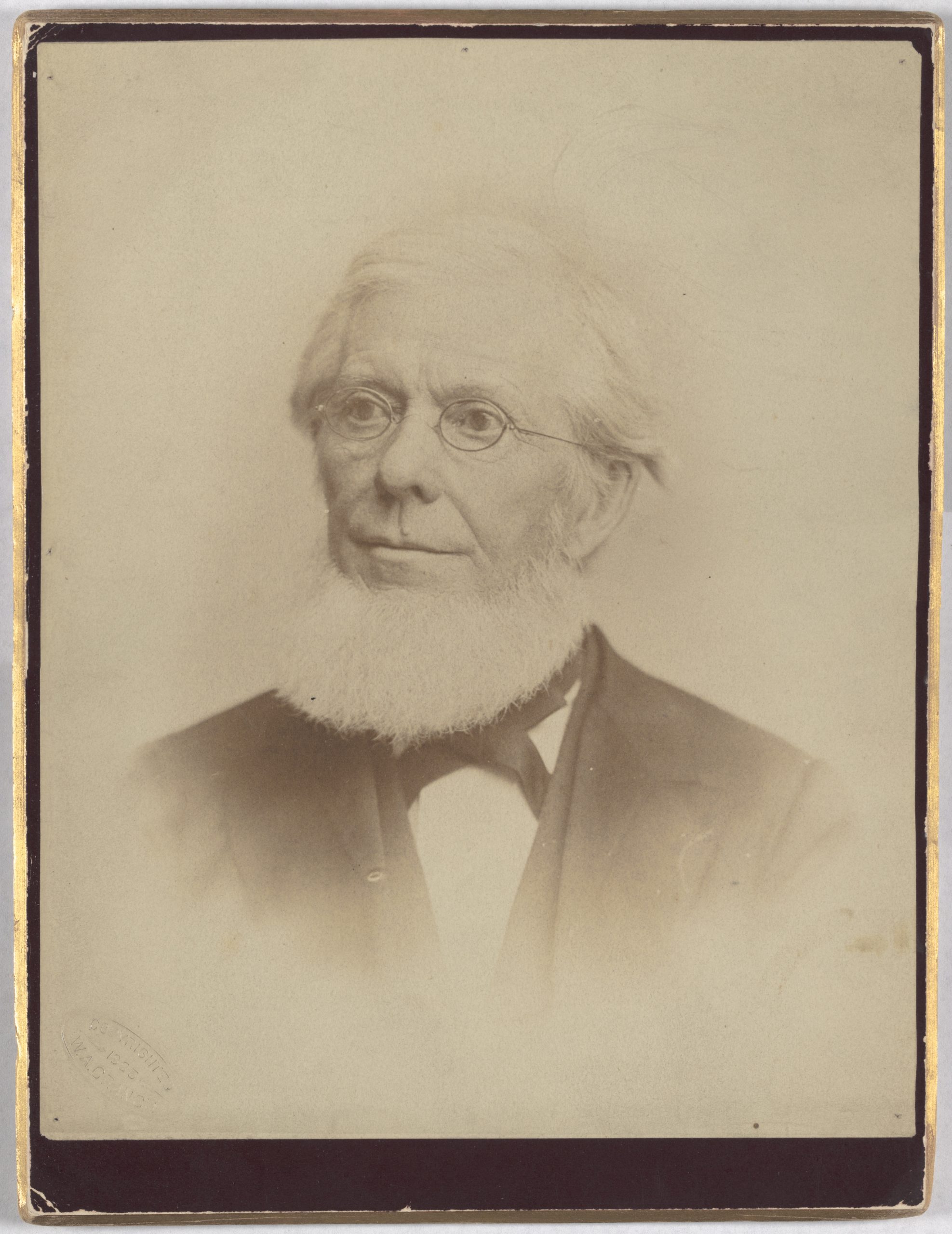 A man smiles at the camera. He wears glasses and has a bowtie. He also has a thick white beard