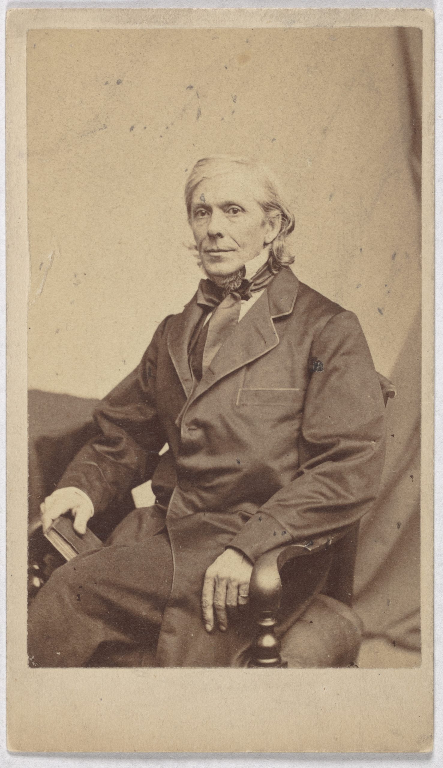 A man with long white hair stares out from where he sits in a chair. He wears a dark colored suit jacket.