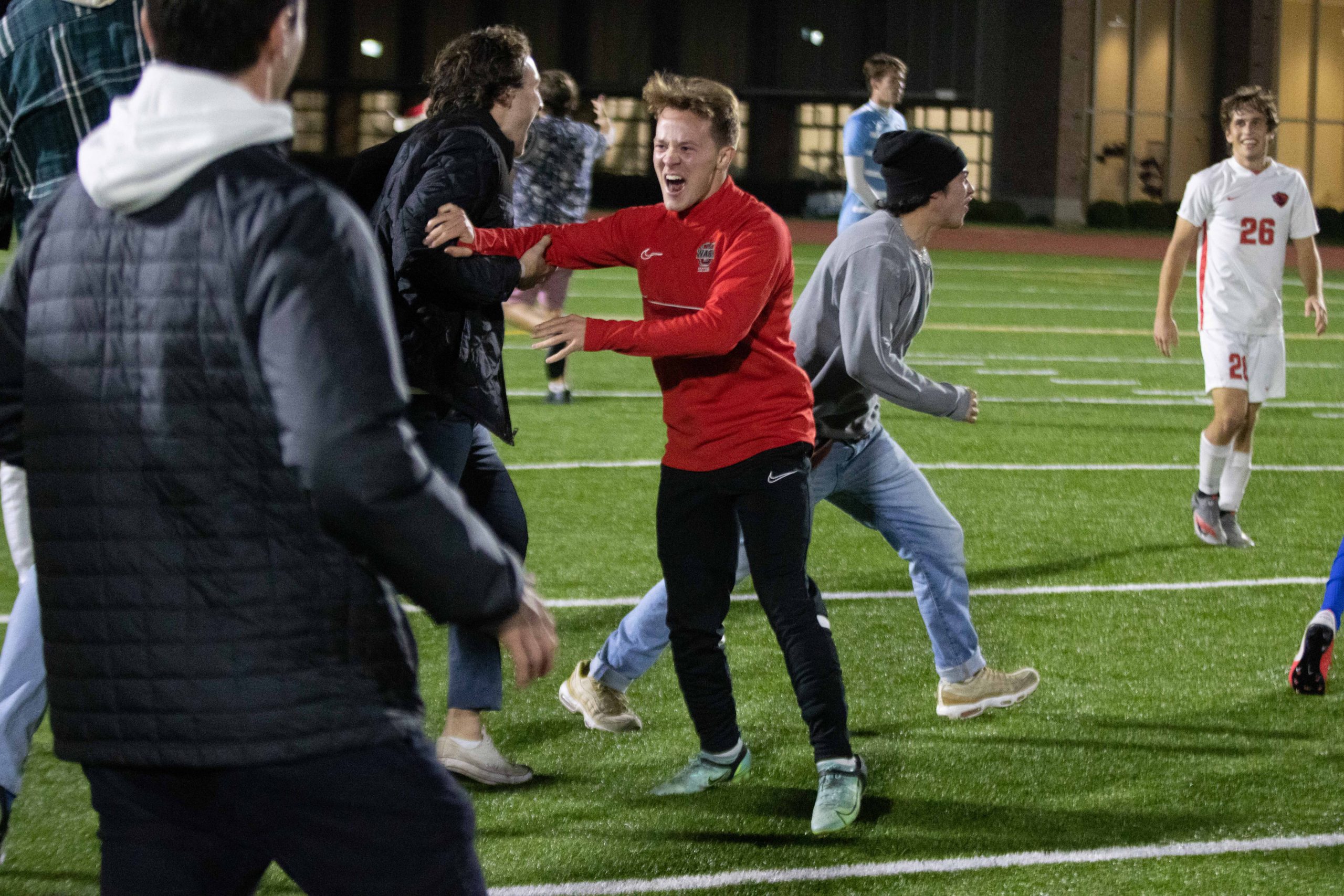 A person in a red shirt and black sweat pants yells happily as other people in non-soccer clothes (jackets and jeans) rush onto the soccer field.