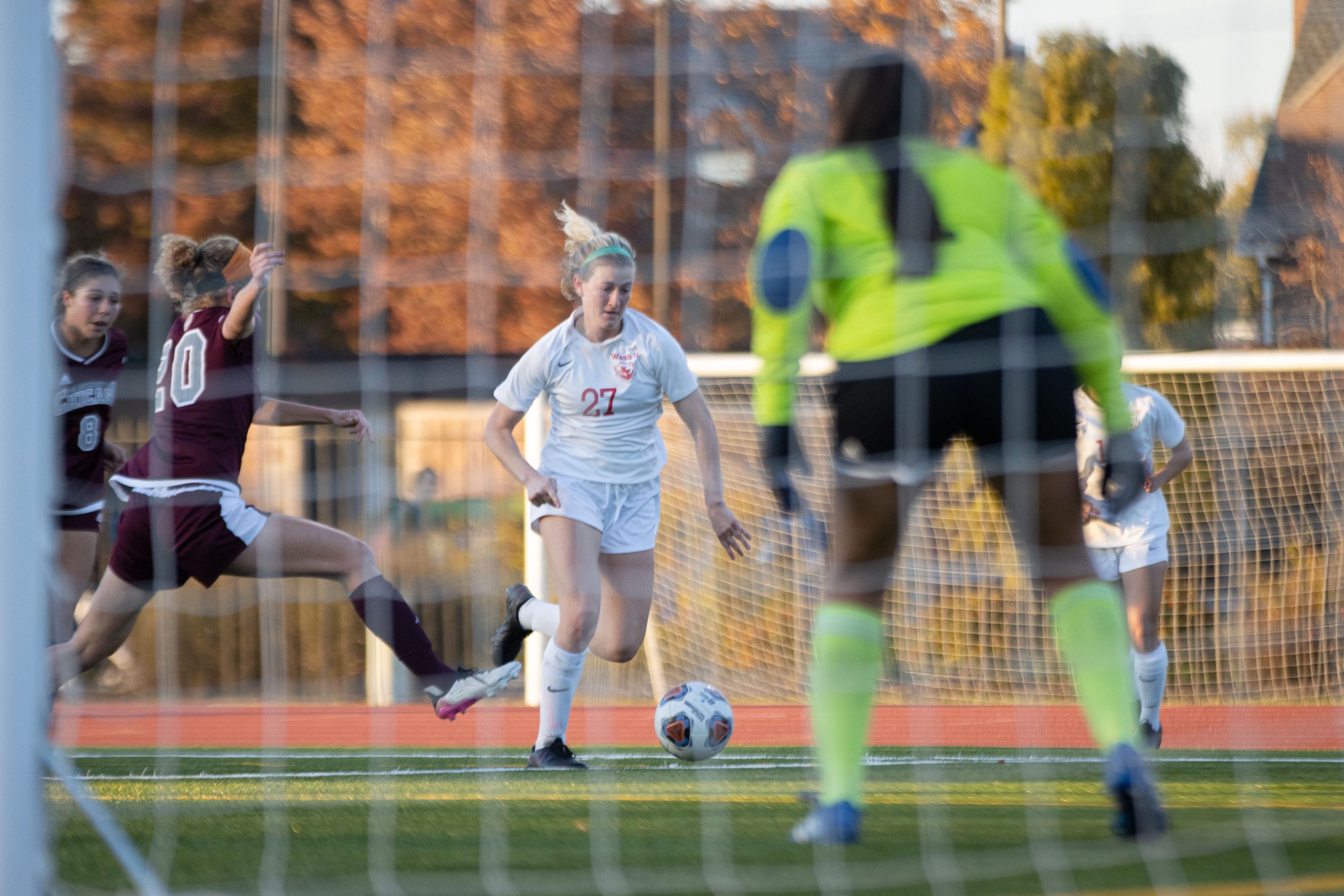 A soccer player in a white jersey wearing the number 27 moves her left leg back to kick the ball toward the goal, where a player wearing a bright neon yelllow shirt stands waiting.