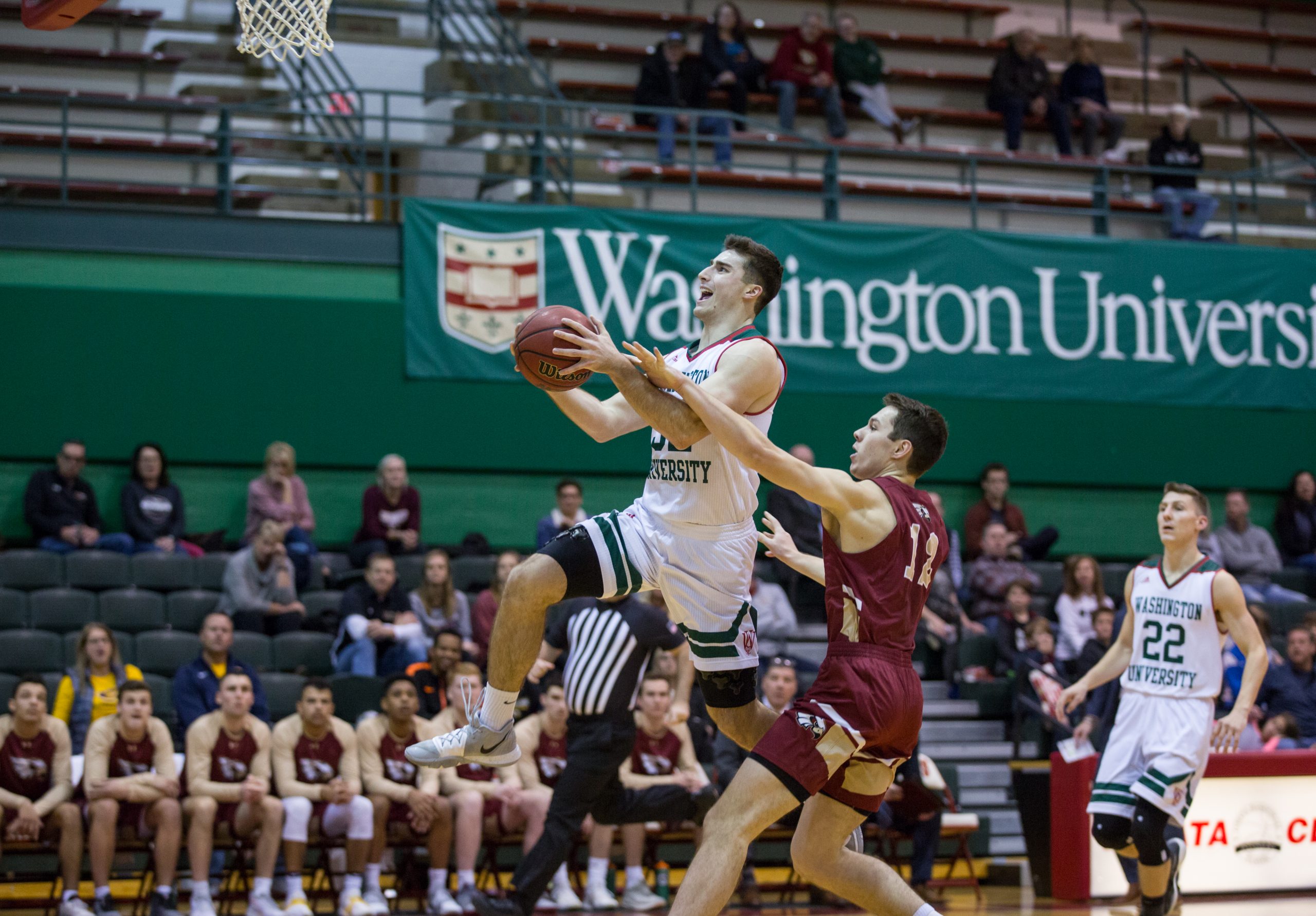 A basketball player in a white uniform clutches a basketball between his two hands as he goes up for a shot while a player in a maroon jersey and the white number 11 sticks his left arm in his way. White letters on a green background read "Washington University" off the court above the stands.