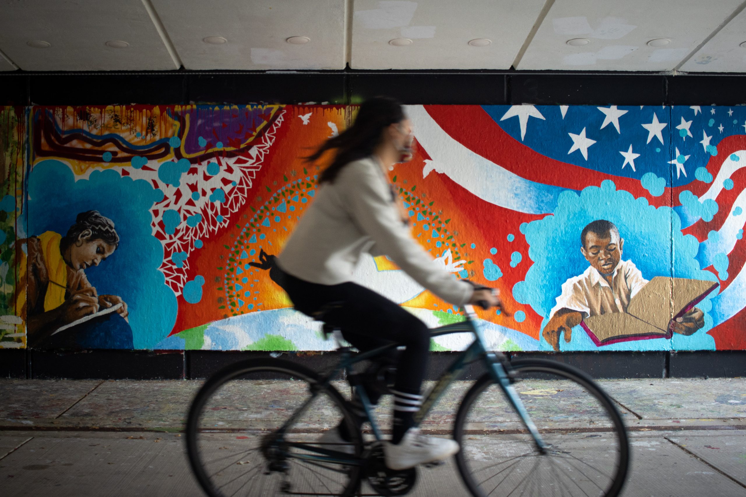 A person in a white shirt and black pants bicycles to the right in front of a colorful mural that displays an orange sunrise, a stylization of the American flag in the upper right hand corner and a man in the lower right hand corner.