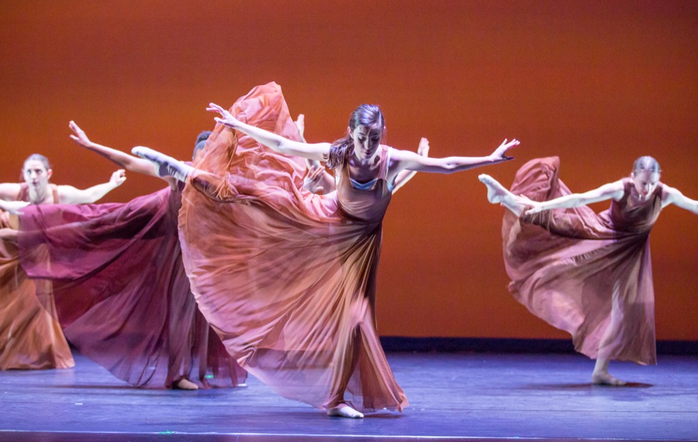 Four dancers in flowy pink dresses take wide bows with their arms spread wide on a stage. 
