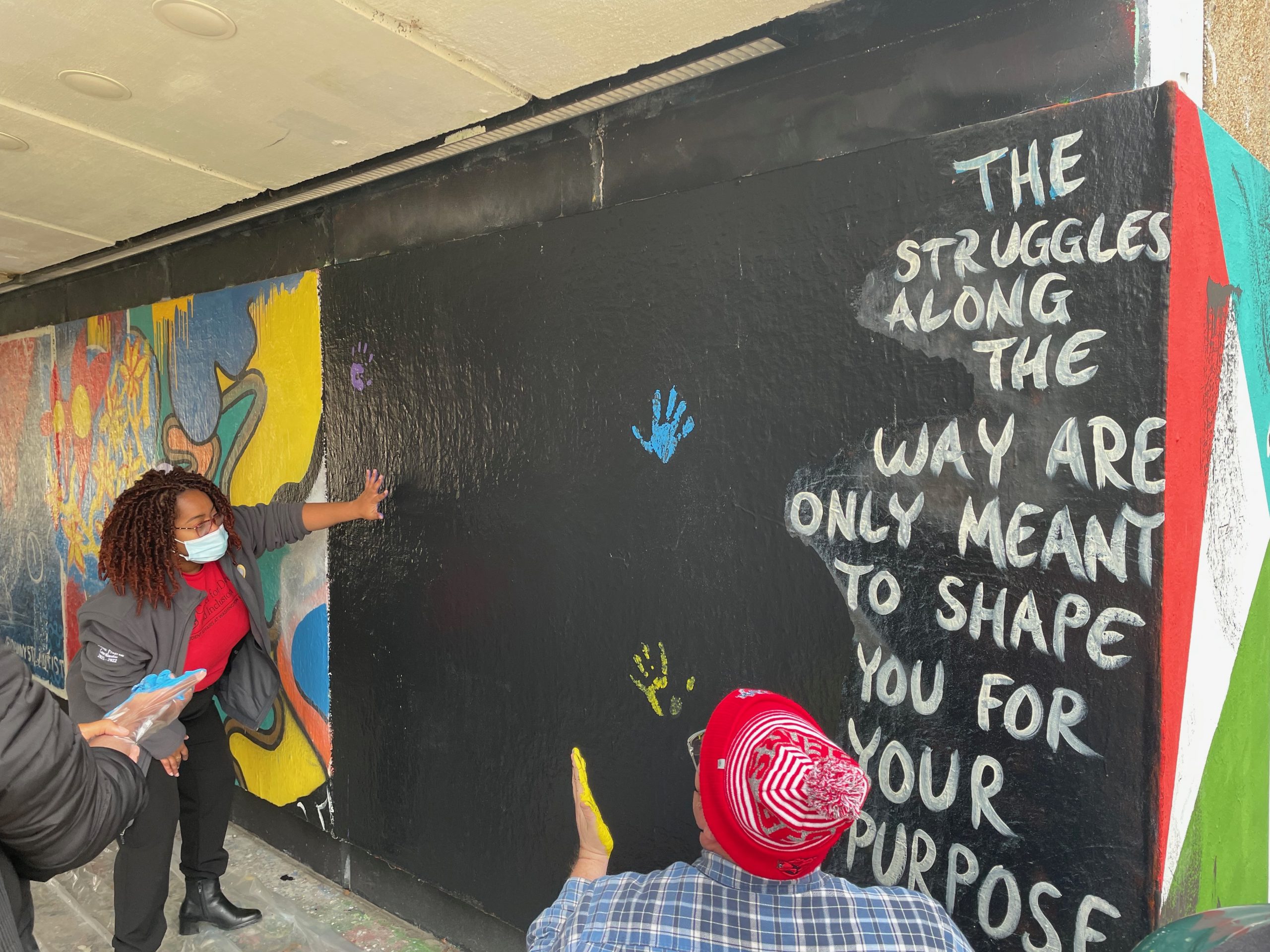 A woman in a gray jacket and red shirt places her hand on a black mural panel, where there are handprints in blue and yellow.