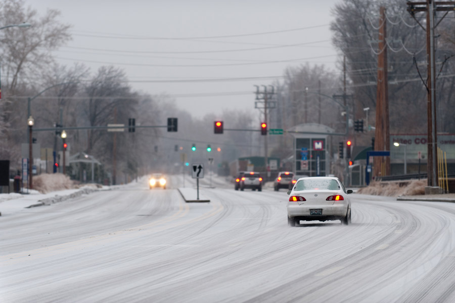 Cars in the snowy right lane of a road wait for a red light as the oncoming lights of another car are seen across the intersection. Green streetlights can be seen in the distance.