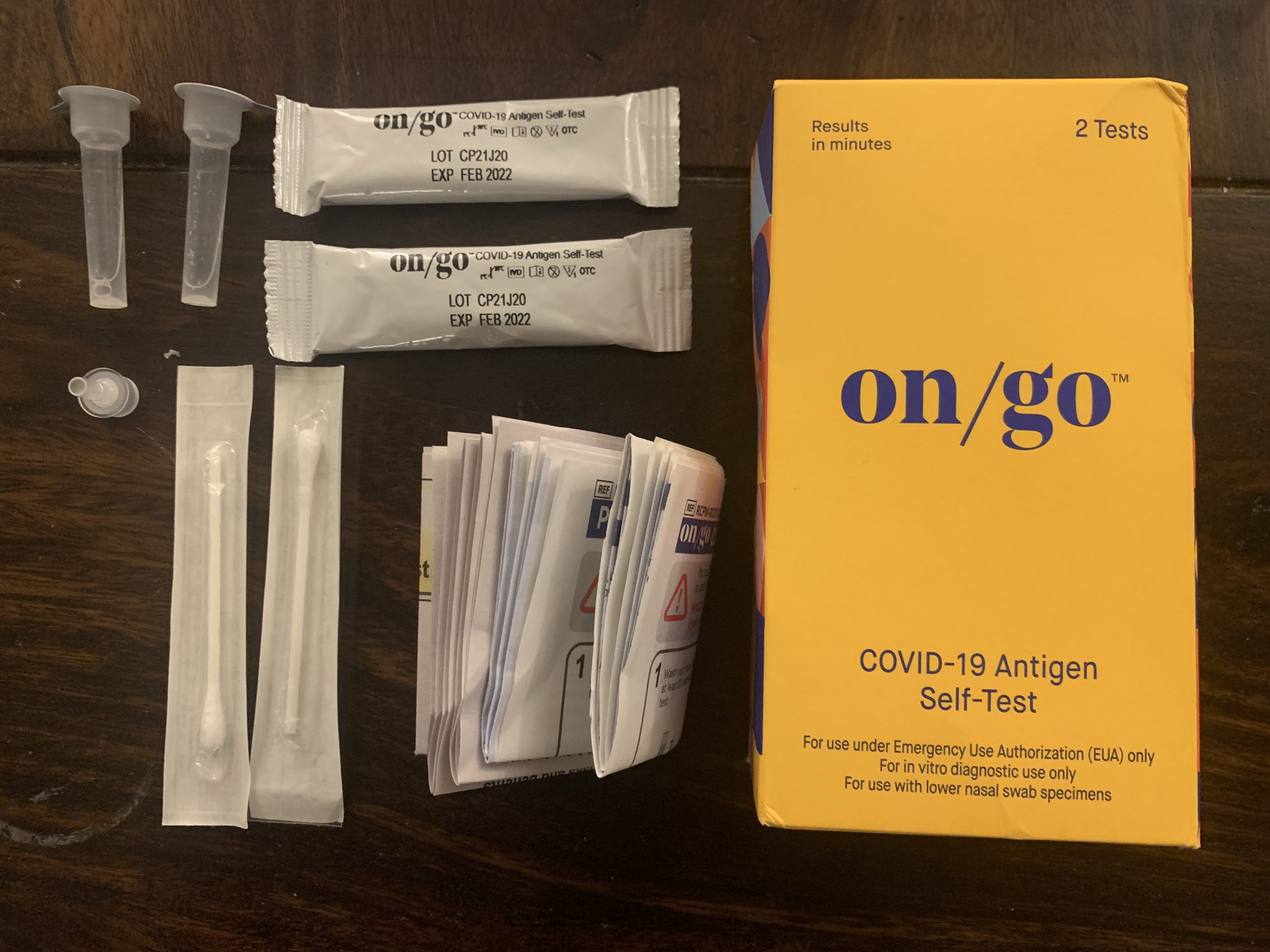 An orange cardboard box sits on a brown wooden surface next to several swabs and test tubes in white packaging
