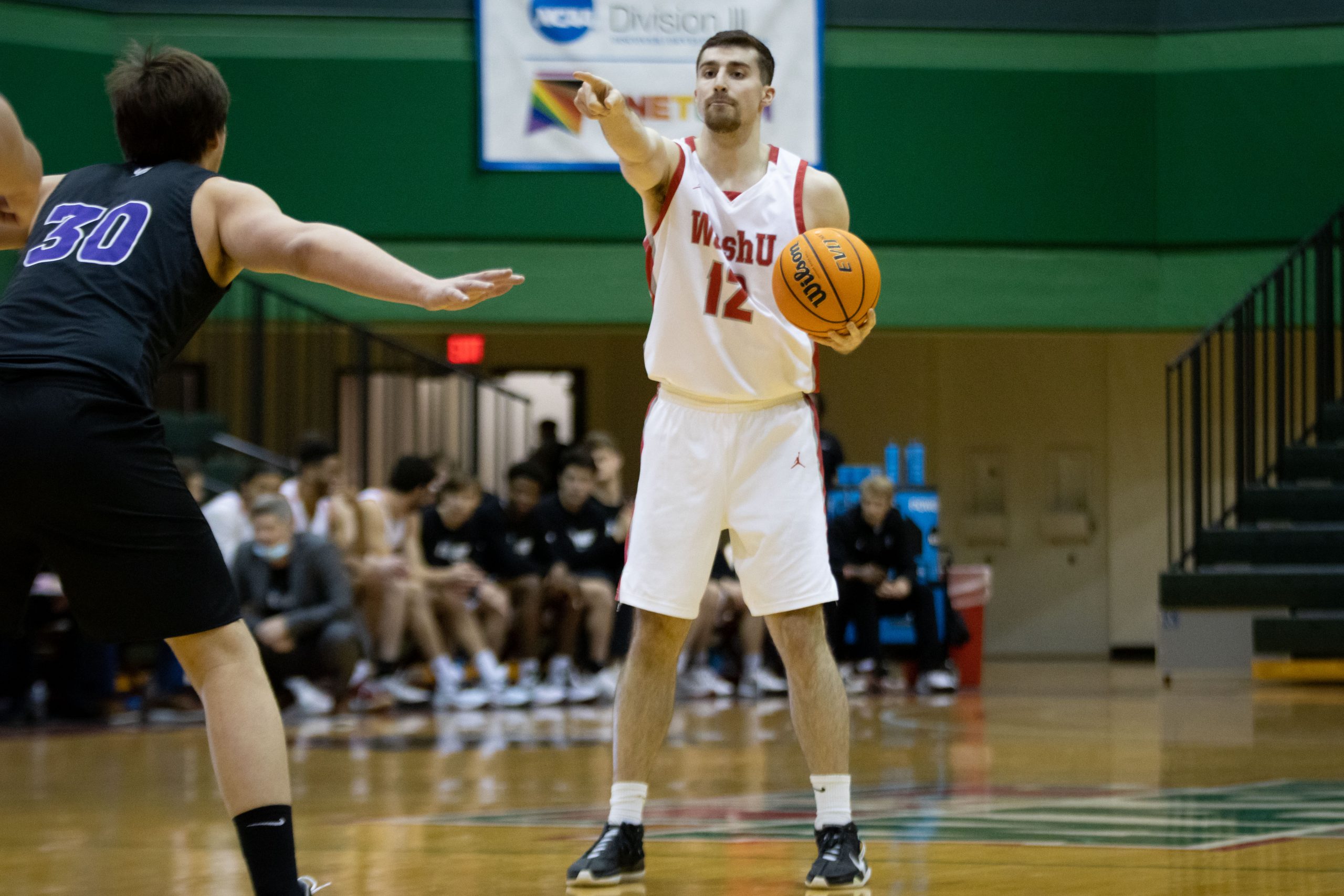 A player in the middle of the frame points with his right hand while holding the ball in his left and wearing a white jersey with red letters that say 'WashU' and the number 12. A defender on the left of the frame with his back to the camera faces the first player in a crouch.
