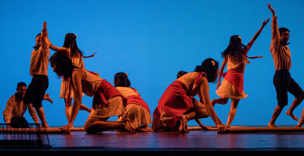 Dancers in white, red and black clothing bend down to grab bamboo poles on a stage, while some on the left and right sides of the stage dance with their hands in the air.