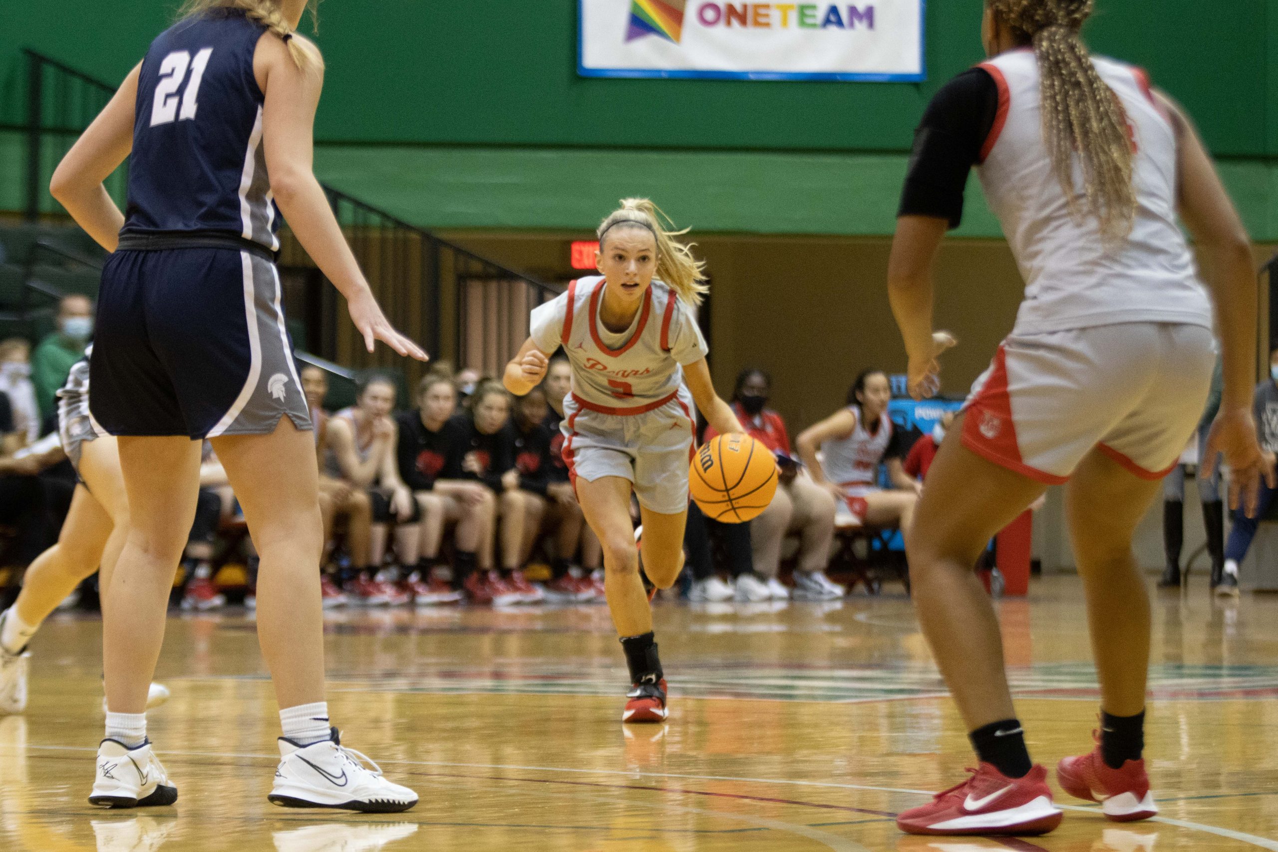 A basketball player dribbles the ball with her left hand as she wears a gray uniform with red trim and stares at a space between a player wearing the same color uniform and one wearing a blue uniform.