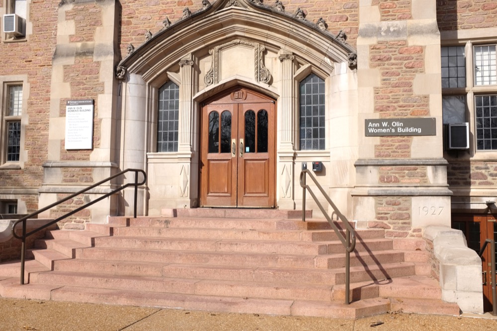 A wooden door stands on a brick facade with steps leading up to it