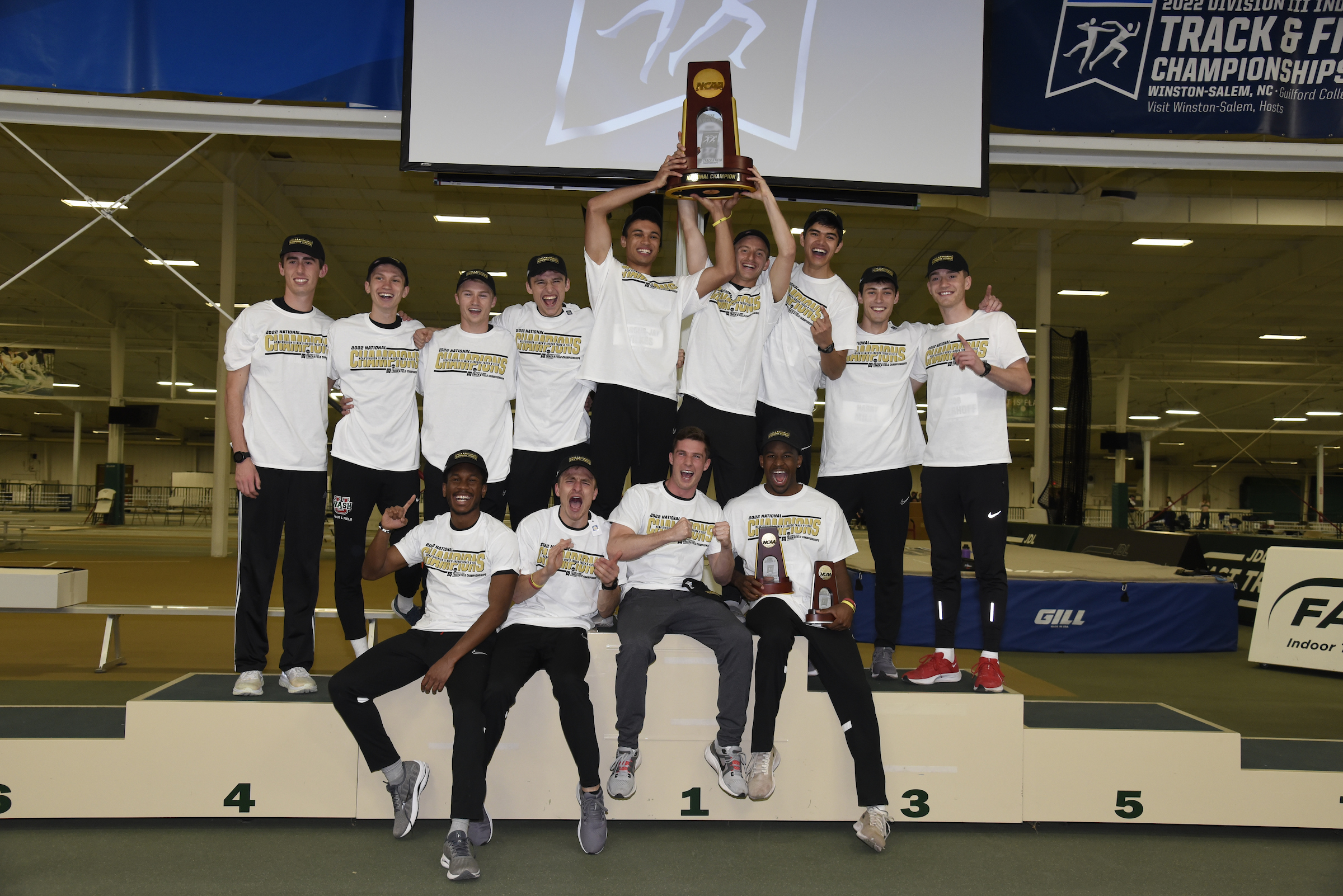 Four young men sit in a row in front of nine others, the middle three of whom hoist a trophy above their heads. The men wear white shirts and black pants. 
