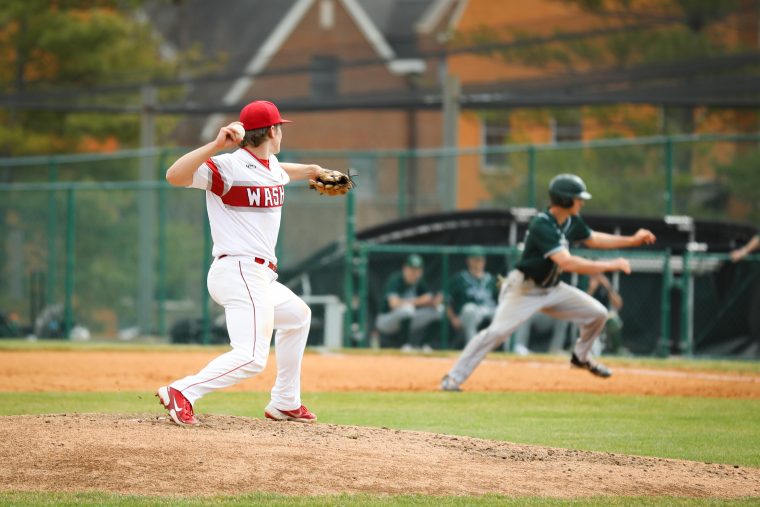 Pitcher in red and white uniform throws the ball to first base; runner, in the background, reverses to try to get to the base. 