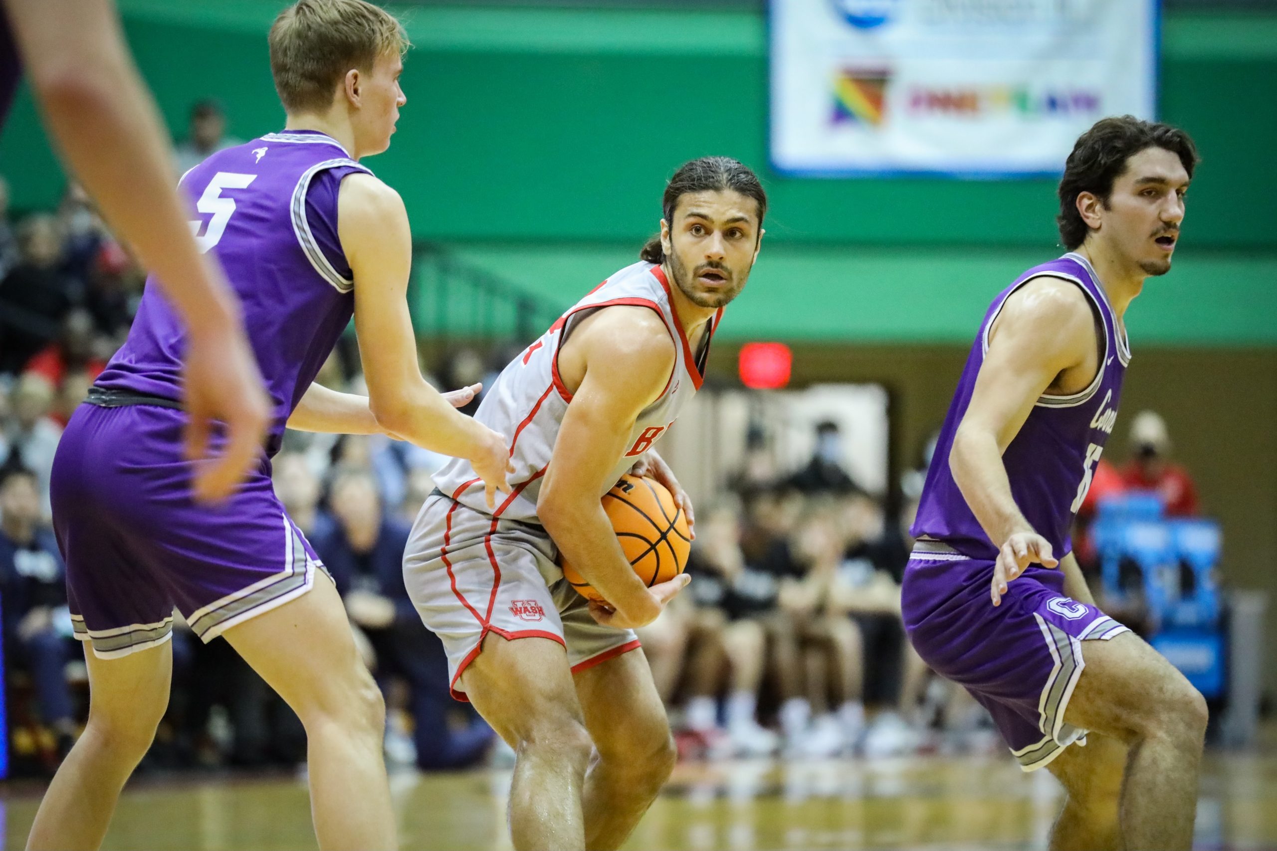 A basketball player in a gray and red jersey holds a ball in two hands close to his stomach to protect it from a defender, who wears a purple jersey and the number 5. Another defender in purple stands on the right side.