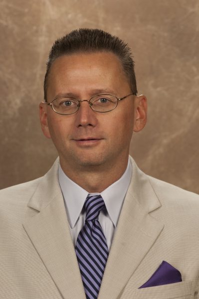 A headshot of Dr. Krister Knapp. He has short brown hair, round glasses, and a blue tie.