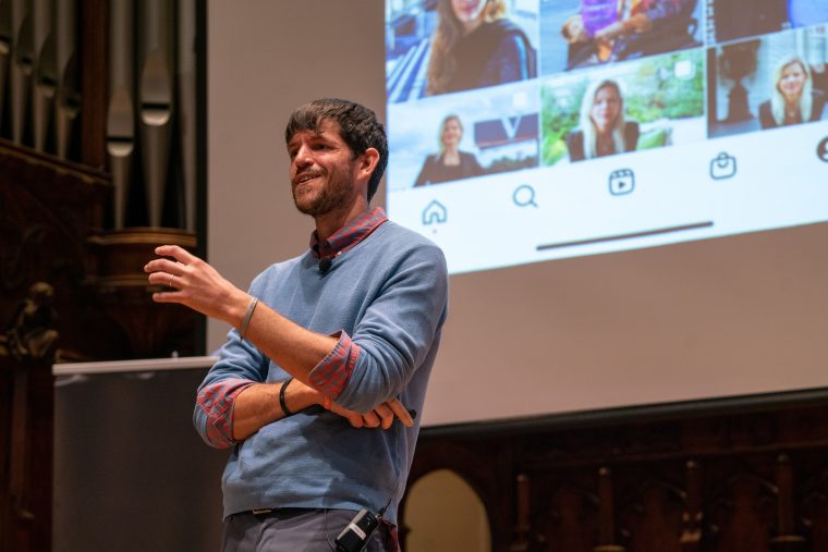 A tall caucasian man with brown hair and a beard wears a blue sweater over a red flannel. He stands in front of a projector showing the Humans of New York page.